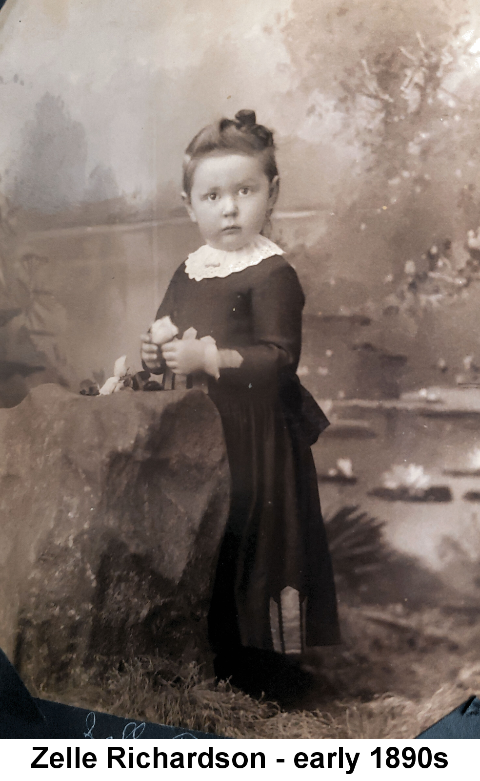 IMAGE/PHOTO: Zelle Richardson - early 1890a: Sepia-tinted full-length photo of a very young girl, hair piled on top of her head, standing on grass just behind a large rock and in front of a painted landscape backdrop, holding a white flower and gazing wide-eyed at the camera.